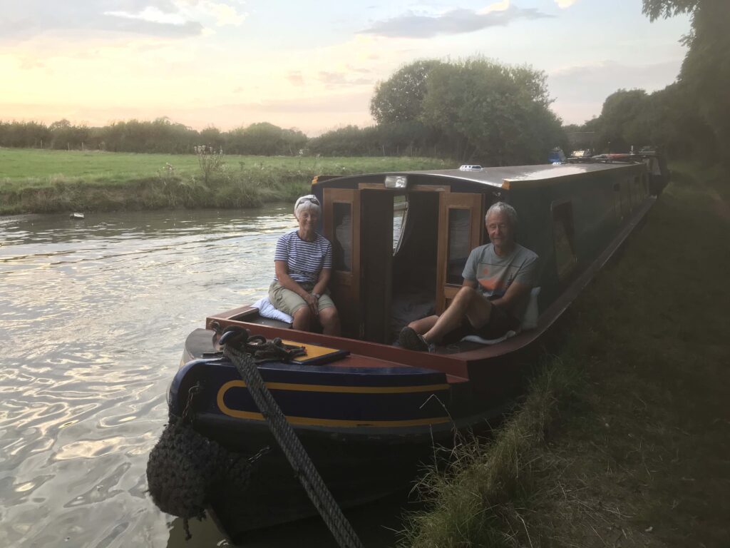 Steve and Leslie are seated in the bow of their hireboat. The sun is setting behind a field and hedgerow on the other side of the canal.