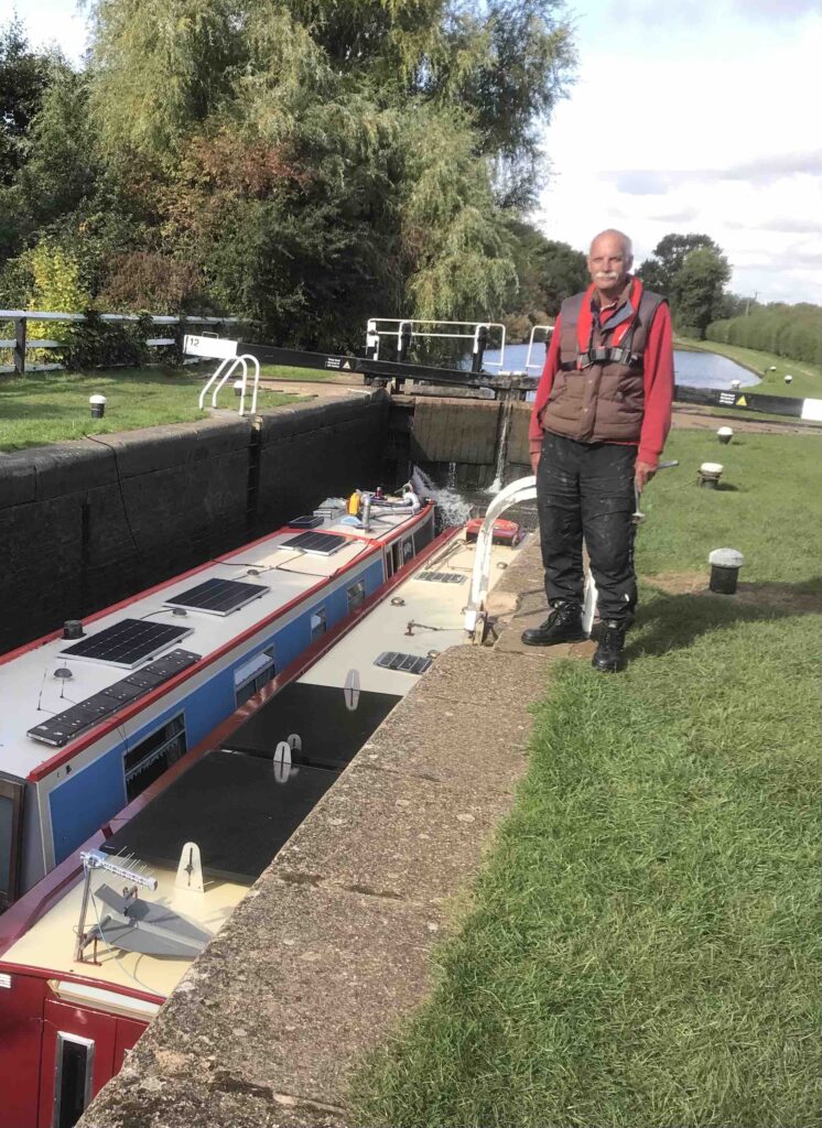 Steve, the retired Military Planner stands at the side of a lock in which his boat and Marry Joan can be seen