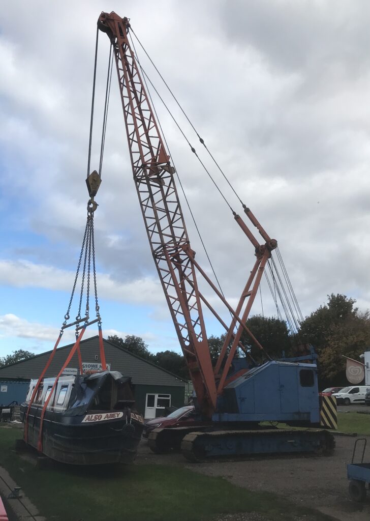 A crane with caterpillar tracks has a cradle hanging from the top of its arm in which a narrowboat is slung