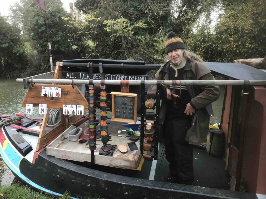 Narrowboater Paul stands in the bows of his boat. He is wearing a bobble hat and a heavy leather overcoat. In front of him are his leather wares he has for sale: coin pouches, bags, wallets, hair grips.
