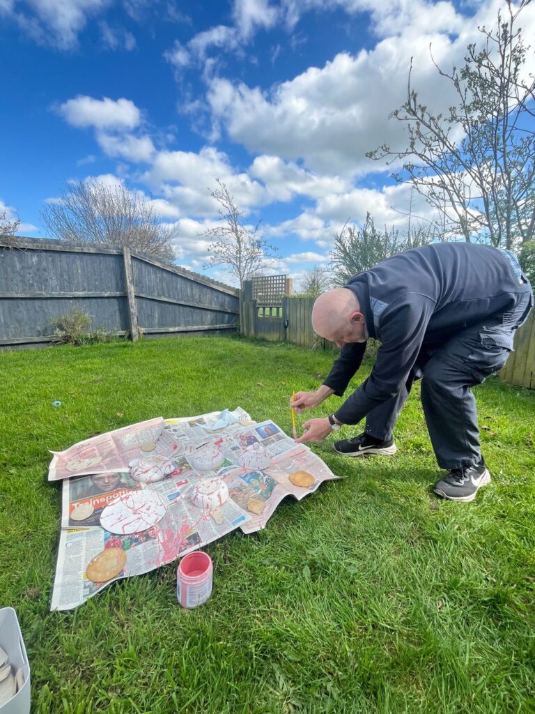 The author is bent over, holding a spatula in front of him. He is in the process of flicking glaze over various items of pottery that are resting on newspaper in a back garden.