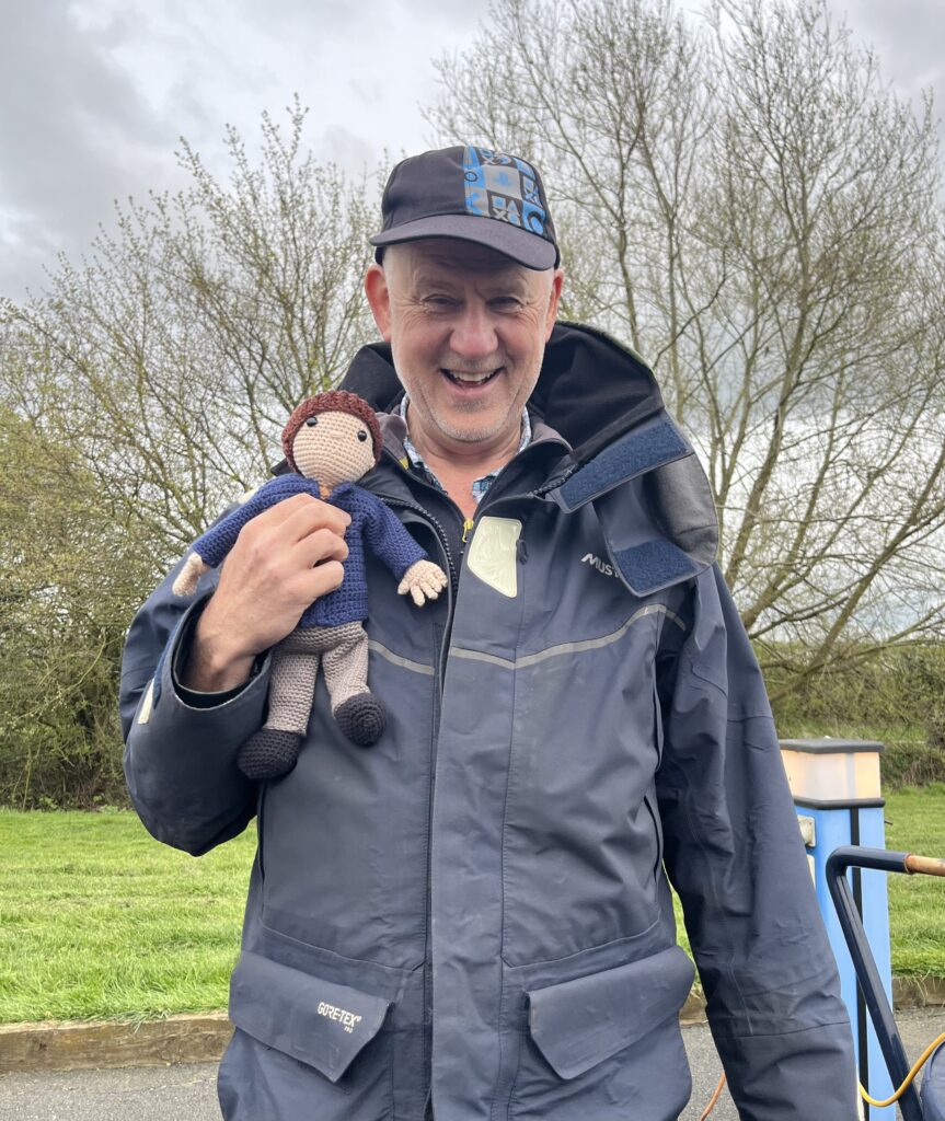The author, who is dressed in a sailing jacket and baseball cap holds a knitted figure in his right hand, against the jacket. The figure has a blue fleece and a brown pom pom hat.